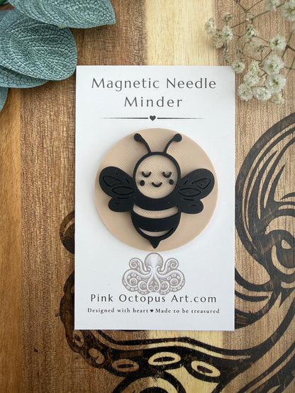 A cute black and beige bee-shaped magnetic needle holder carded on a white 'Magnetic Needle Minder' display card by Pink Octopus Art.com, styled on a wooden surface with dried flowers.