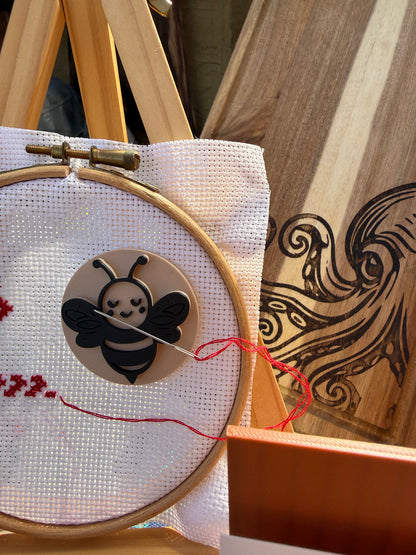 A close-up of a wooden embroidery hoop holding white fabric with a wooden bee applique and red thread, alongside a wooden board with engraved black wave-like patterns.