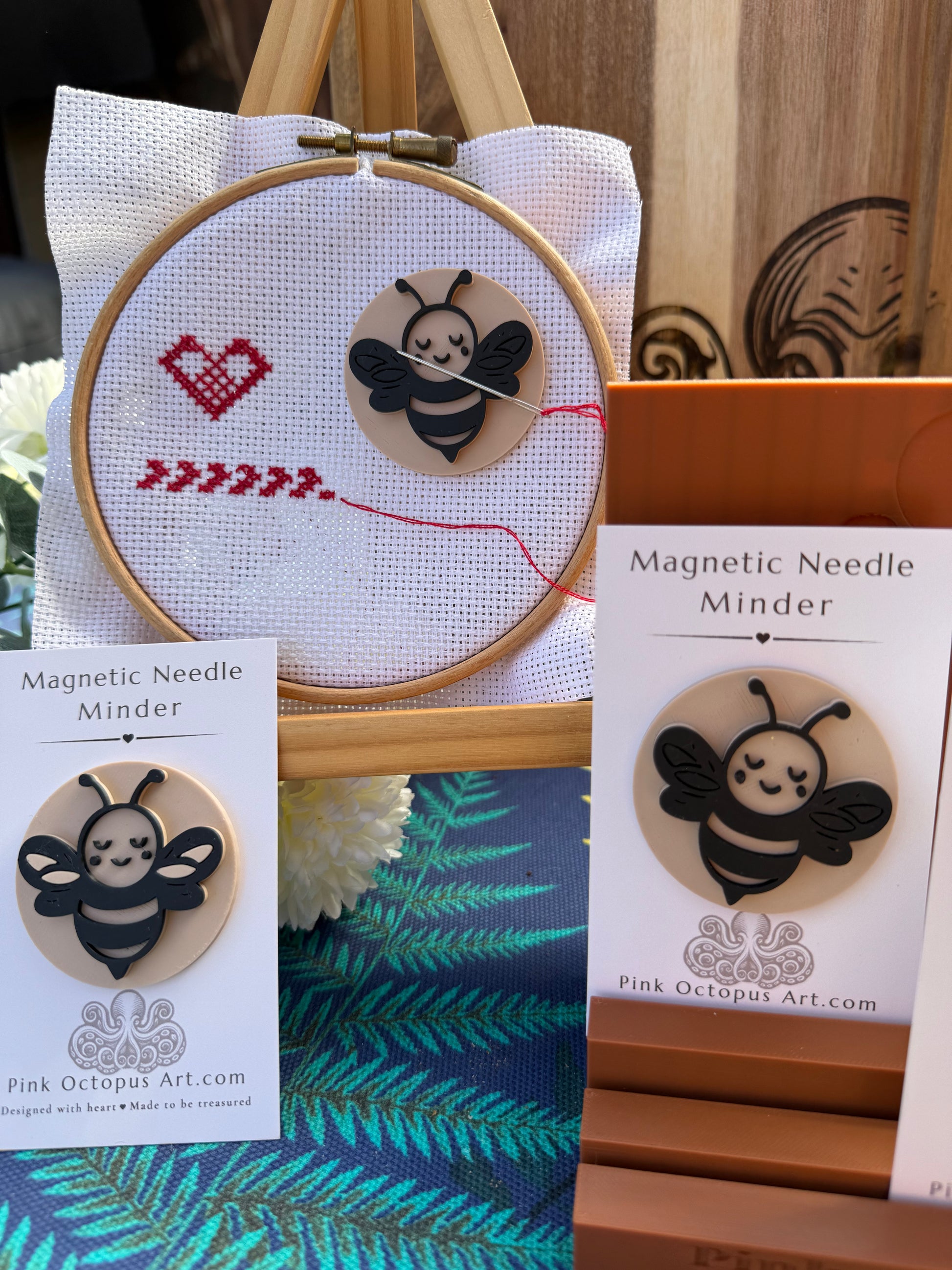 A cute cartoon bee magnetic needle minder displayed in a craft setting, shown both mounted on a wooden embroidery hoop with red thread and cross-stitch details, and on retail display cards next to its orange packaging box. The brand 'Pink Octopus Art' is visible on the cards.