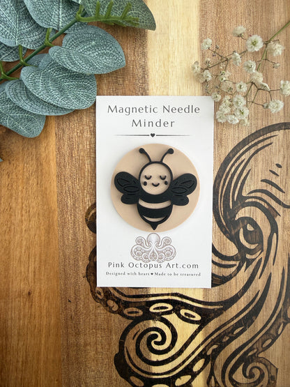 A decorative card displaying a bee-shaped magnetic needle holder labeled 'Magnetic Needle Minder' and 'Pink Octopus Art.com,' styled on a wooden surface with green leaves and small white flowers.