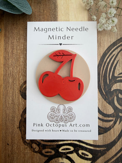 A red cherry-shaped magnetic needle minder displayed on its white backing card, which reads 'Magnetic Needle Minder' and shows the brand 'Pink Octopus Art.com', set against a rustic wooden background with dried flowers.