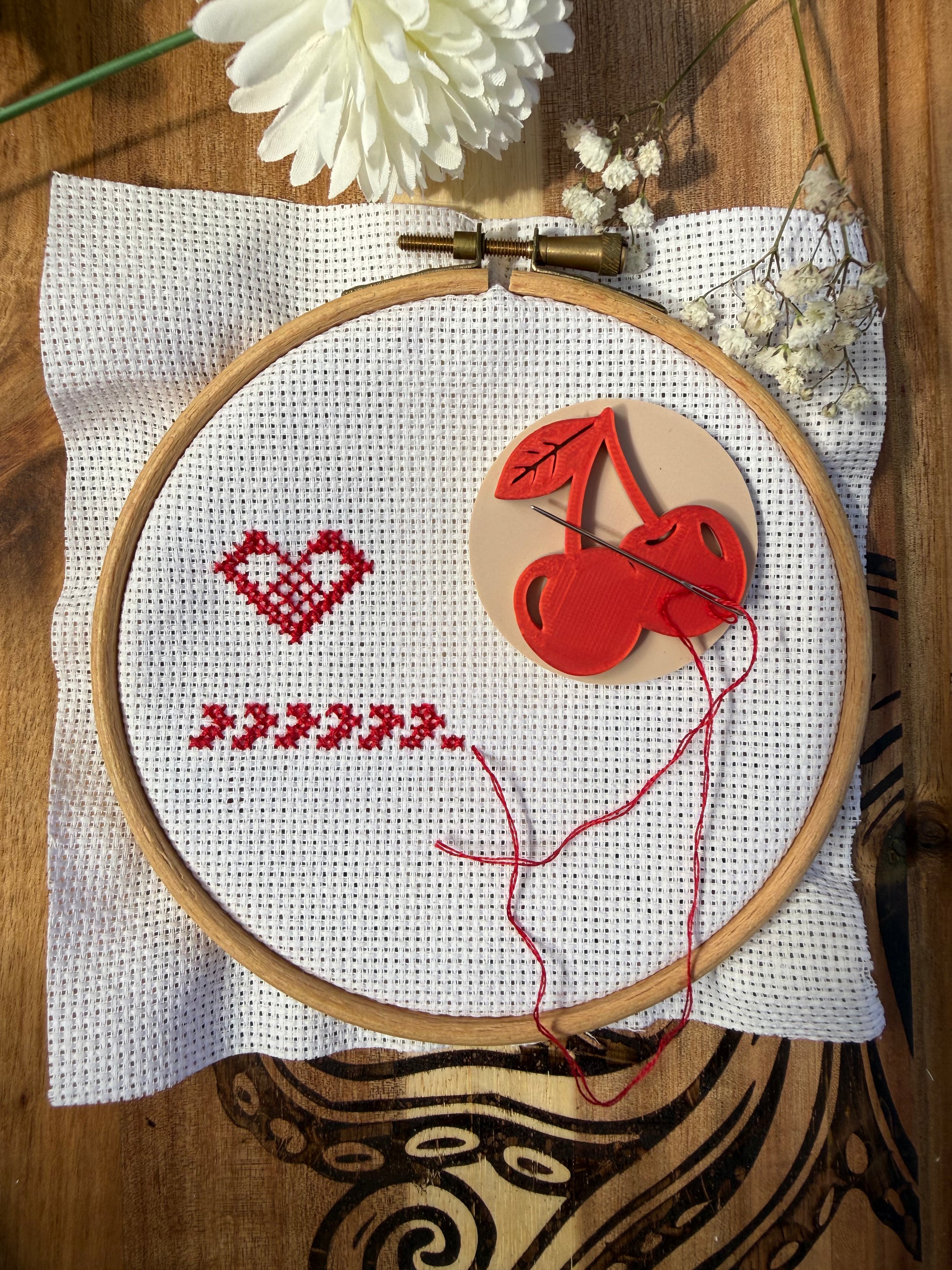 A wooden embroidery hoop holding white A4 fabric with a red stitched heart and chevron pattern, alongside a red cherry magnetic needle minder and thread. The hoop is placed on a white cloth over a dark wood surface, octopus  with dried flowers and a white flower in the background.