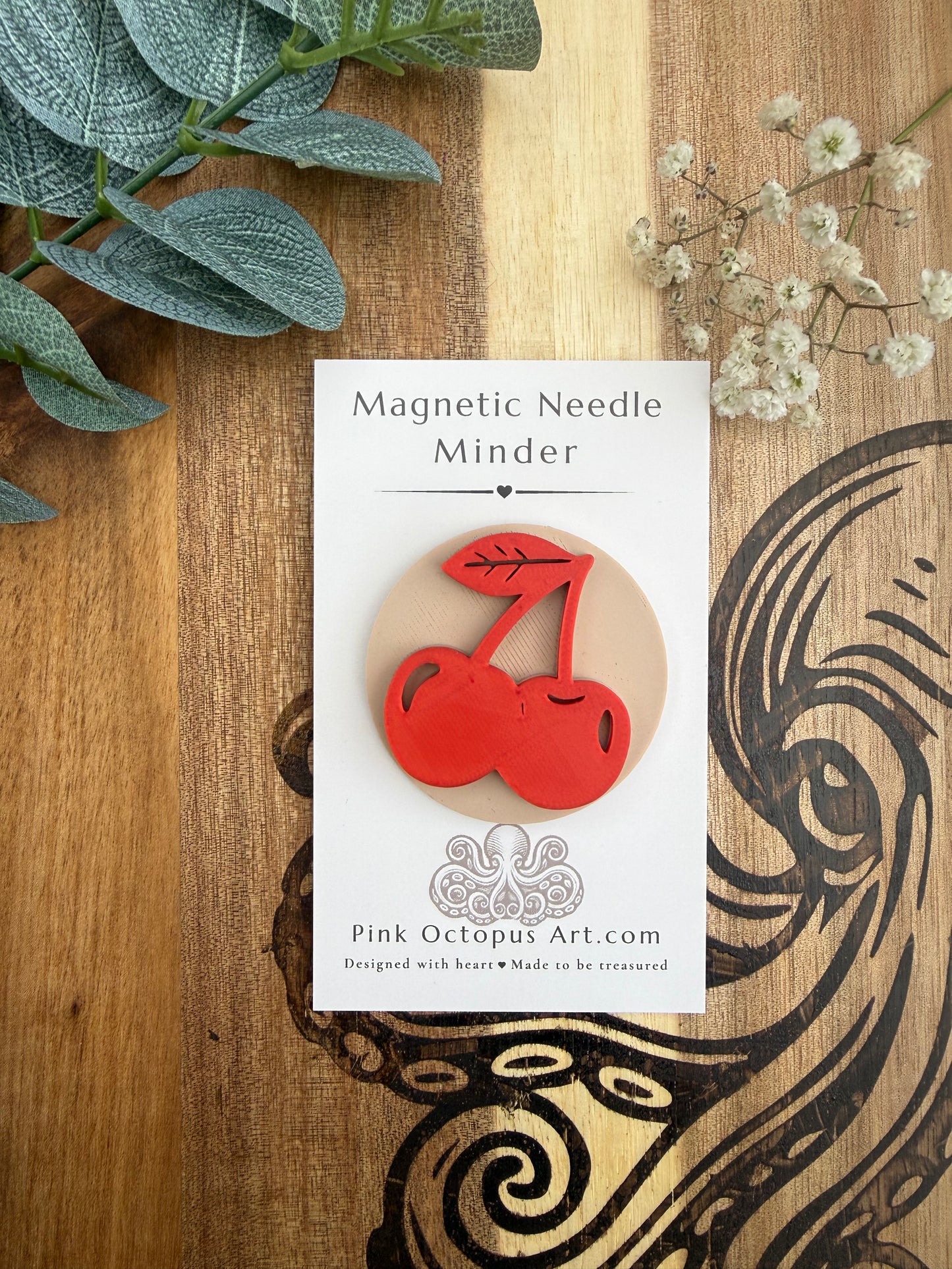 A cherry-shaped magnetic needle pin by Pink Octopus Art is displayed on its white backing card, which is placed on a wooden surface decorated with green leaves and small white flowers.