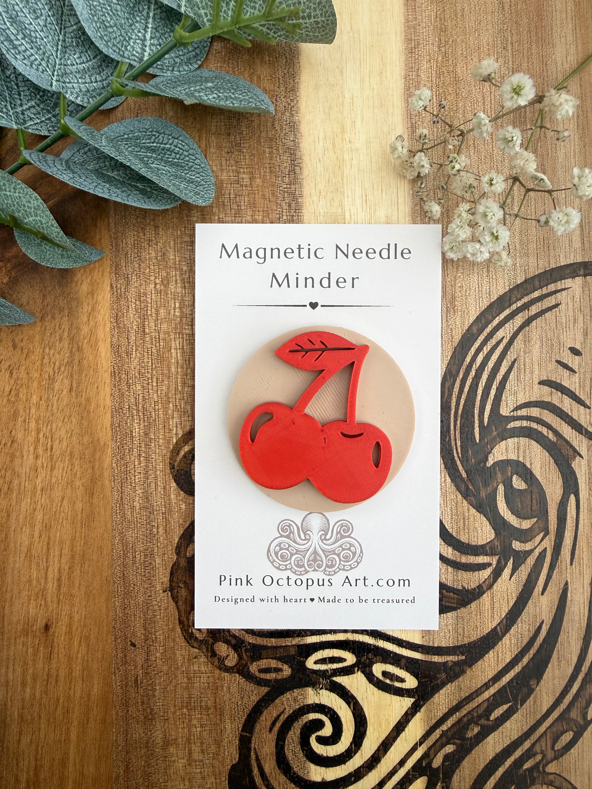 A cherry-shaped magnetic needle pin by Pink Octopus Art is displayed on its white backing card, which is placed on a wooden surface decorated with green leaves and small white flowers.