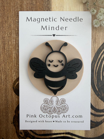 A cute black and beige magnetic bee ornament mounted on a white backing card labeled 'Magnetic Needle Minder' with 'Pink Octopus Art.com' printed below, photographed on a wood surface.