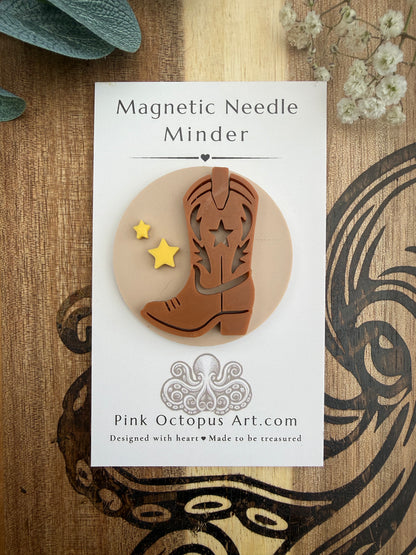 A magnetic needle minder shaped like a brown cowboy boot with two small yellow star cutouts, mounted on a white backing card labeled 'Magnetic Needle Minder' and 'Pink Octopus Art.com,' photographed on a wood surface with decorative dried flowers and a teal leaf.