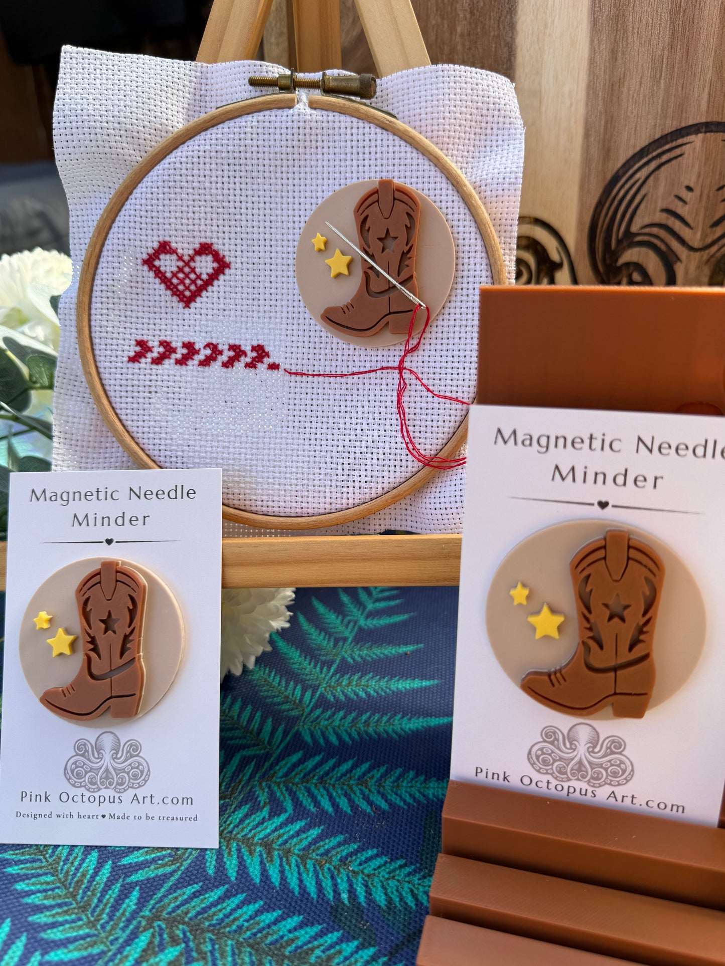 A handmade wooden cowboy boot magnet with embroidered stars and stripes, displayed alongside its packaging and a demonstration of a magnetic needle in an embroidery hoop.