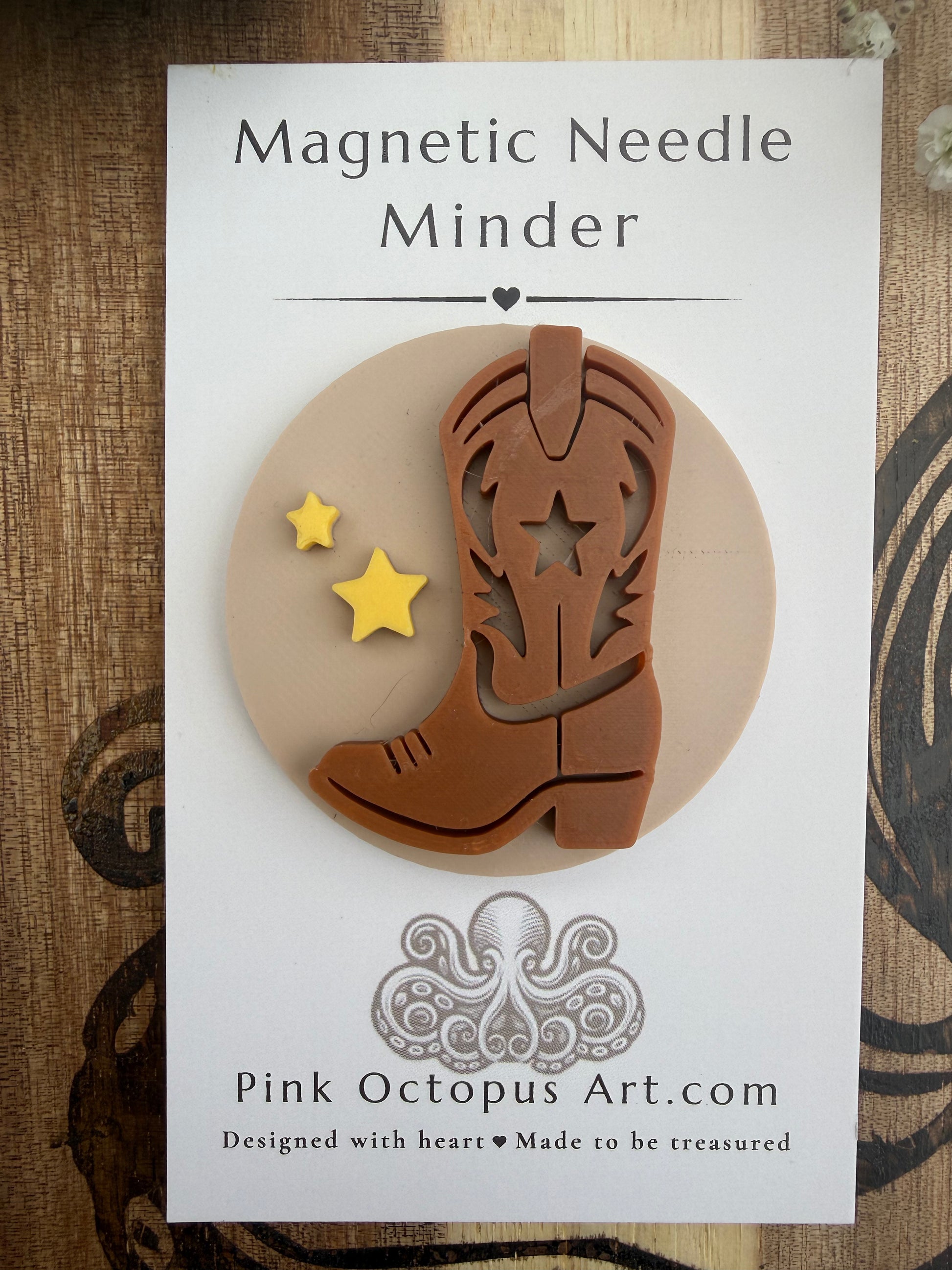 A brown cowboy boot-shaped magnetic needle minder with star cutouts and two small yellow stars, mounted on a white backing card labeled 'Magnetic Needle Minder' by Pink Octopus Art.com, photographed on a wood surface.