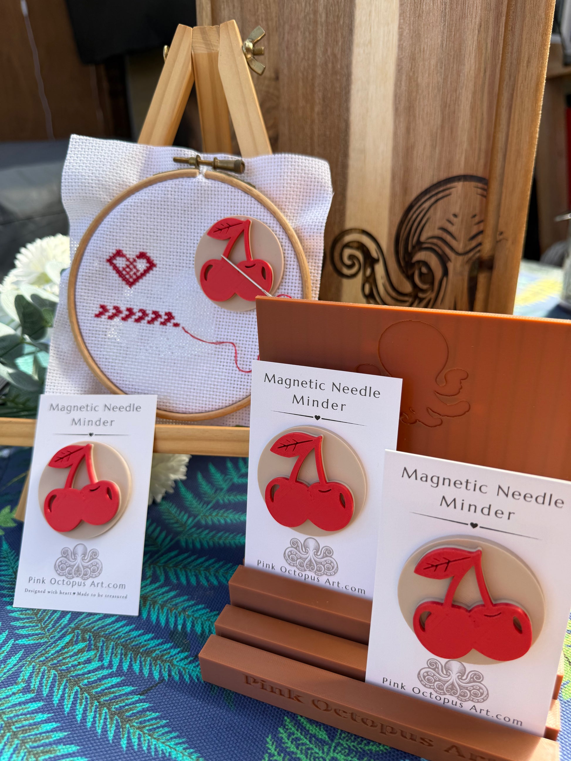 Three 'Magnetic Needle Minder' craft kits by Pink Octopus Art are displayed on wooden stands, alongside an embroidery hoop with a cherry design on white fabric.