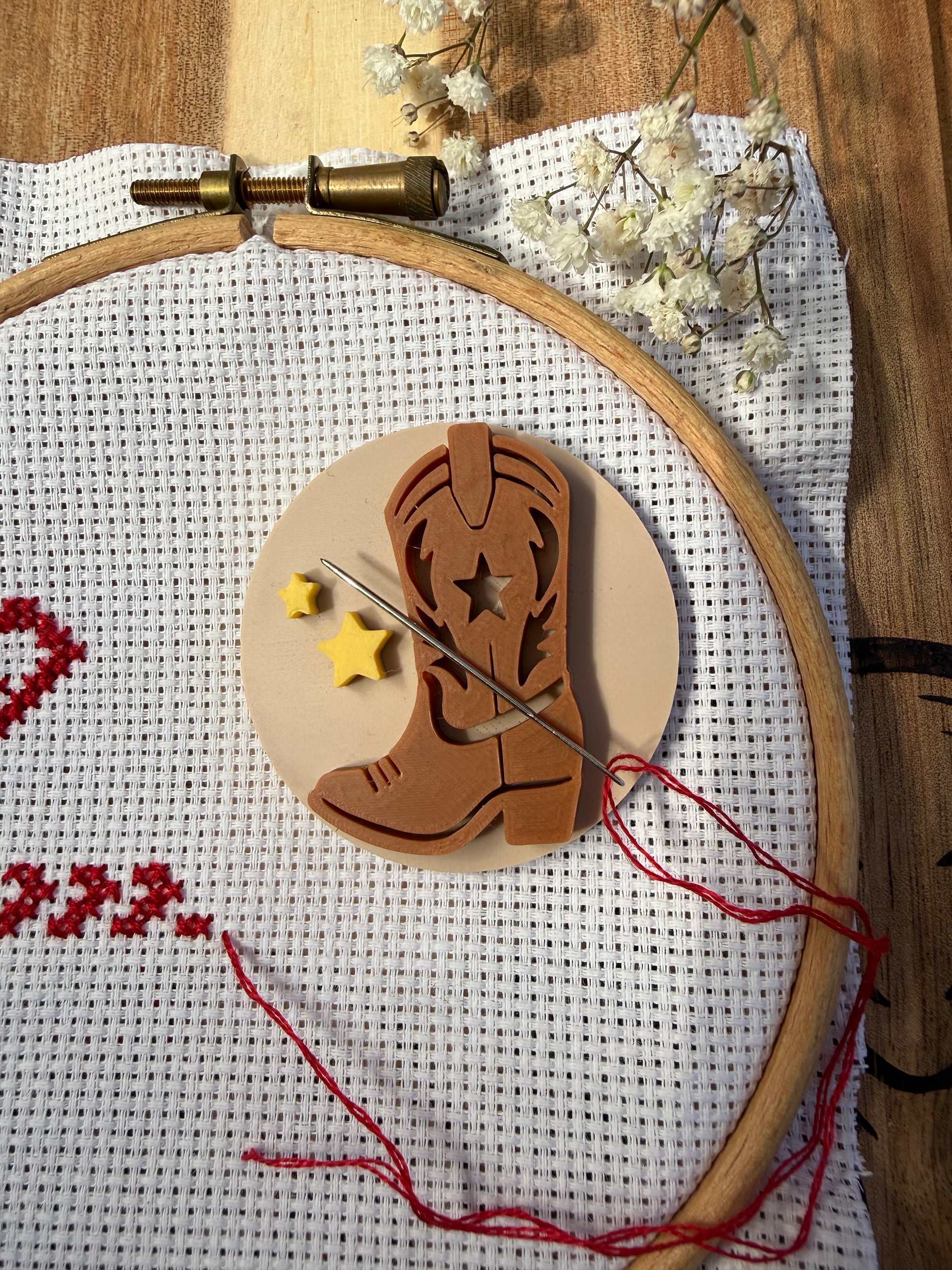 A wooden cowboy boot-shaped sewing or embroidery tool with a star cutout, displayed on a wooden hoop over white cross-stitch fabric with red stitching, thread, a needle, and small white flowers.