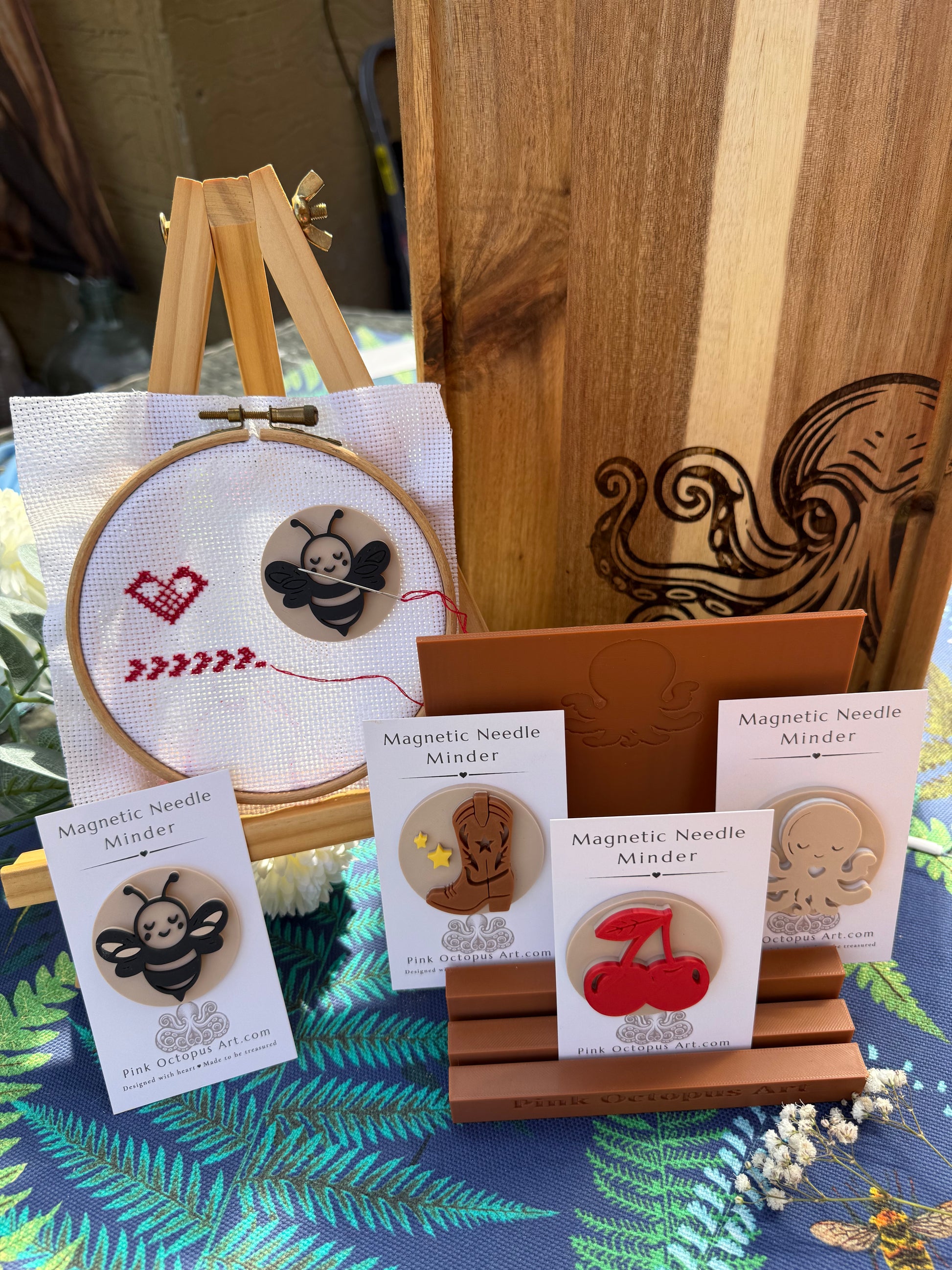 A display of wooden magnetic needle pincushions by Pink Octopus Art., featuring various designs like a bee, cherries, and a cowboy boot on display cards, a wooden stand, and a sample embroidery hoop showing a bee design.