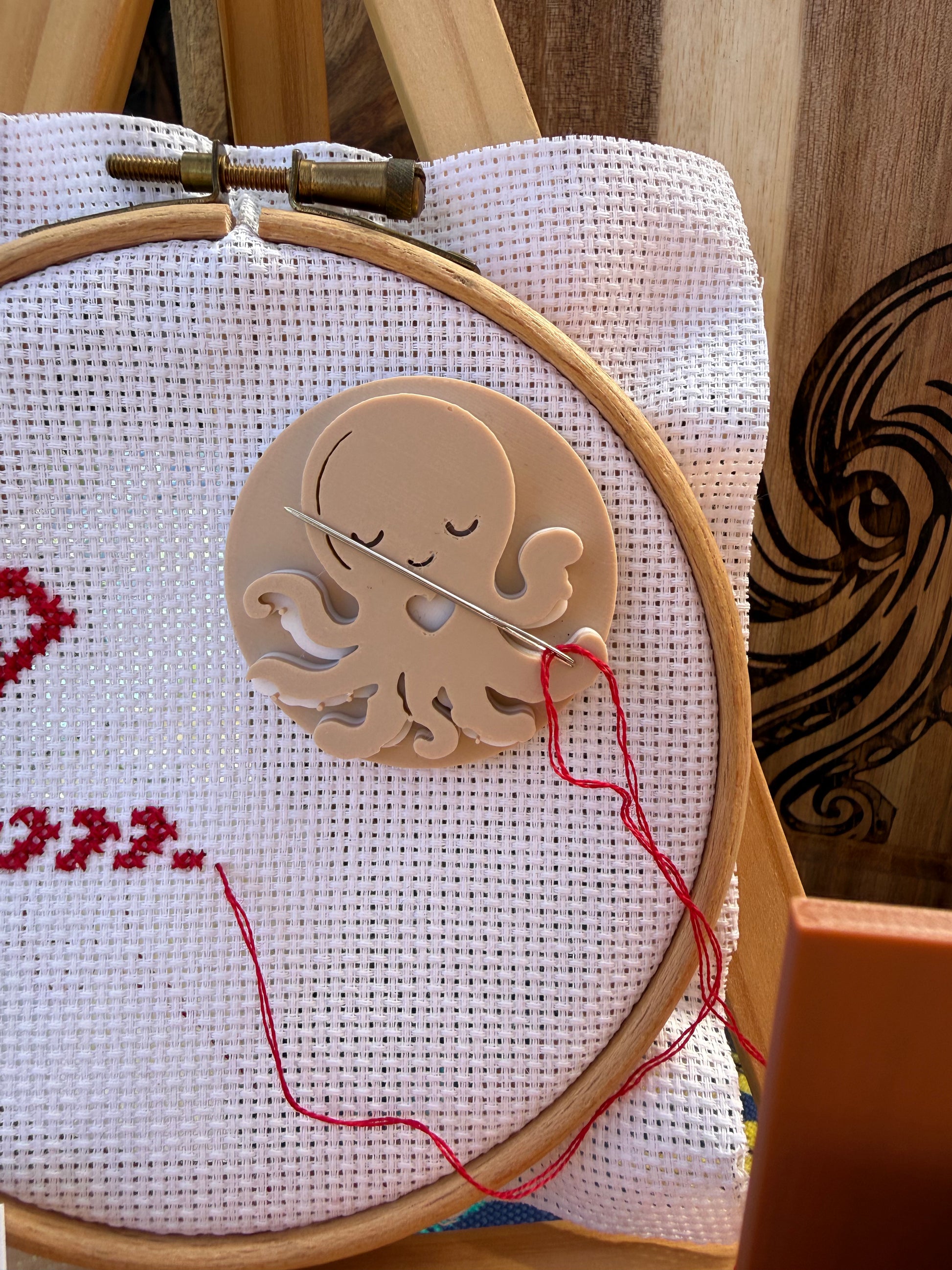 A close-up of a wooden octopus-shaped needle holder with a wooden needle resting inside it, mounted on white cross-stitch fabric inside a wooden embroidery hoop. Red thread loops down from the octopus.