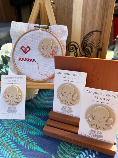 Magnetic Needle Minder wooden craft kit by Pink October Art, featuring a wooden octopus magnet and a wooden embroidery hoop with a white fabric and red stitched heart, displayed on a wooden stand with other product cards.