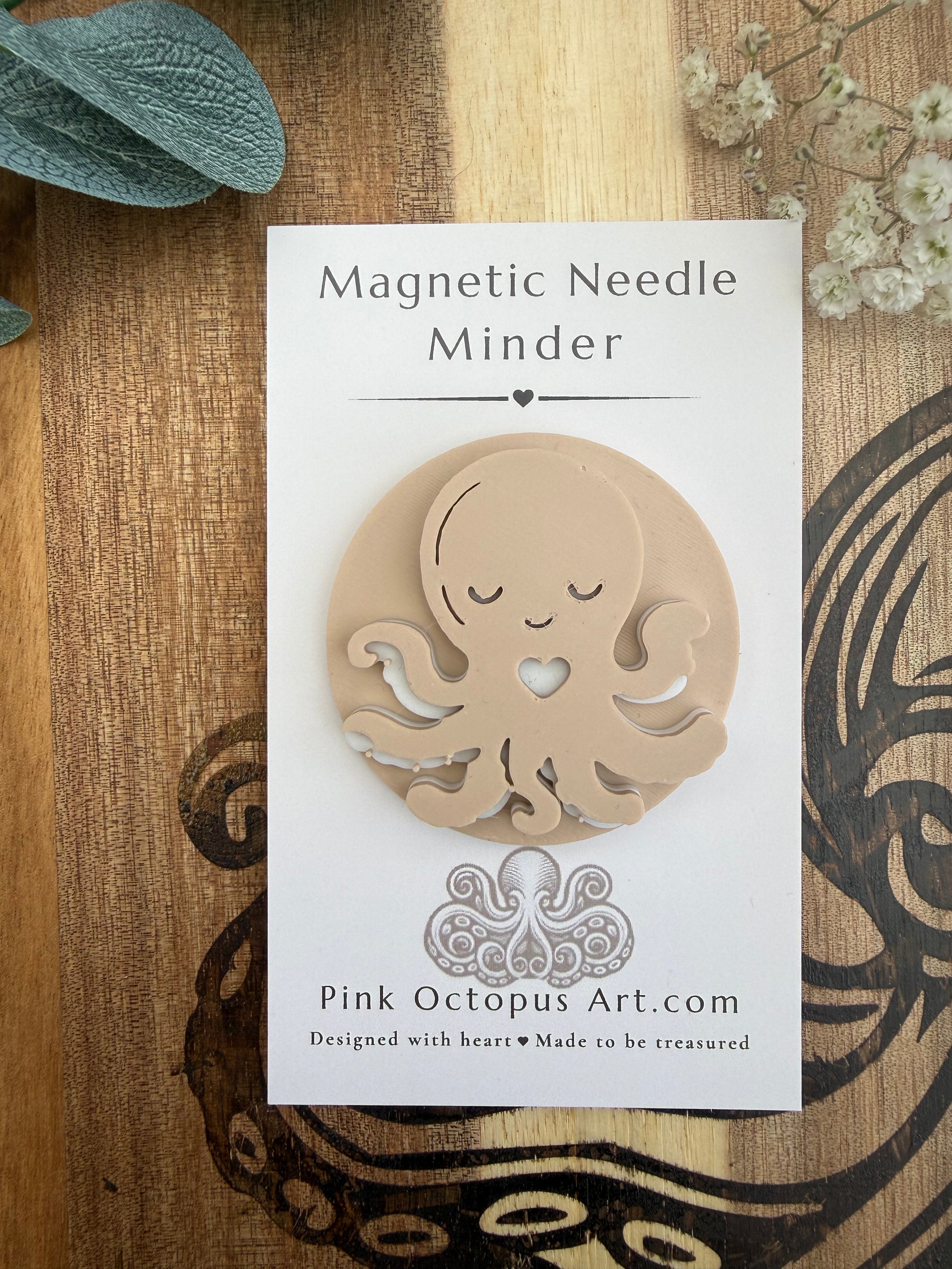 A magnetic needle minder shaped like a cute octopus inside a circle, mounted on its white backing card. The card is displayed on a rustic wooden background with decorative dried flowers and a sage leaf.
