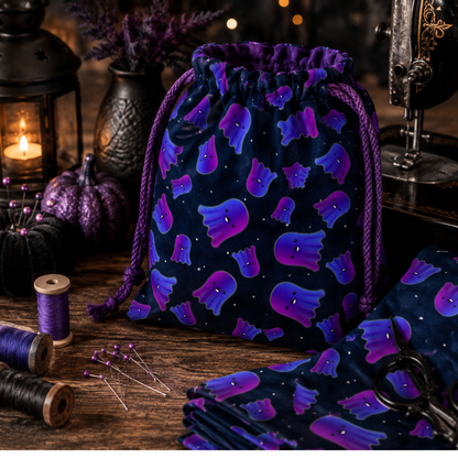 A dark navy drawstring bag with a purple and blue ghost pattern, styled on a rustic wooden table with sewing and Halloween-themed props.