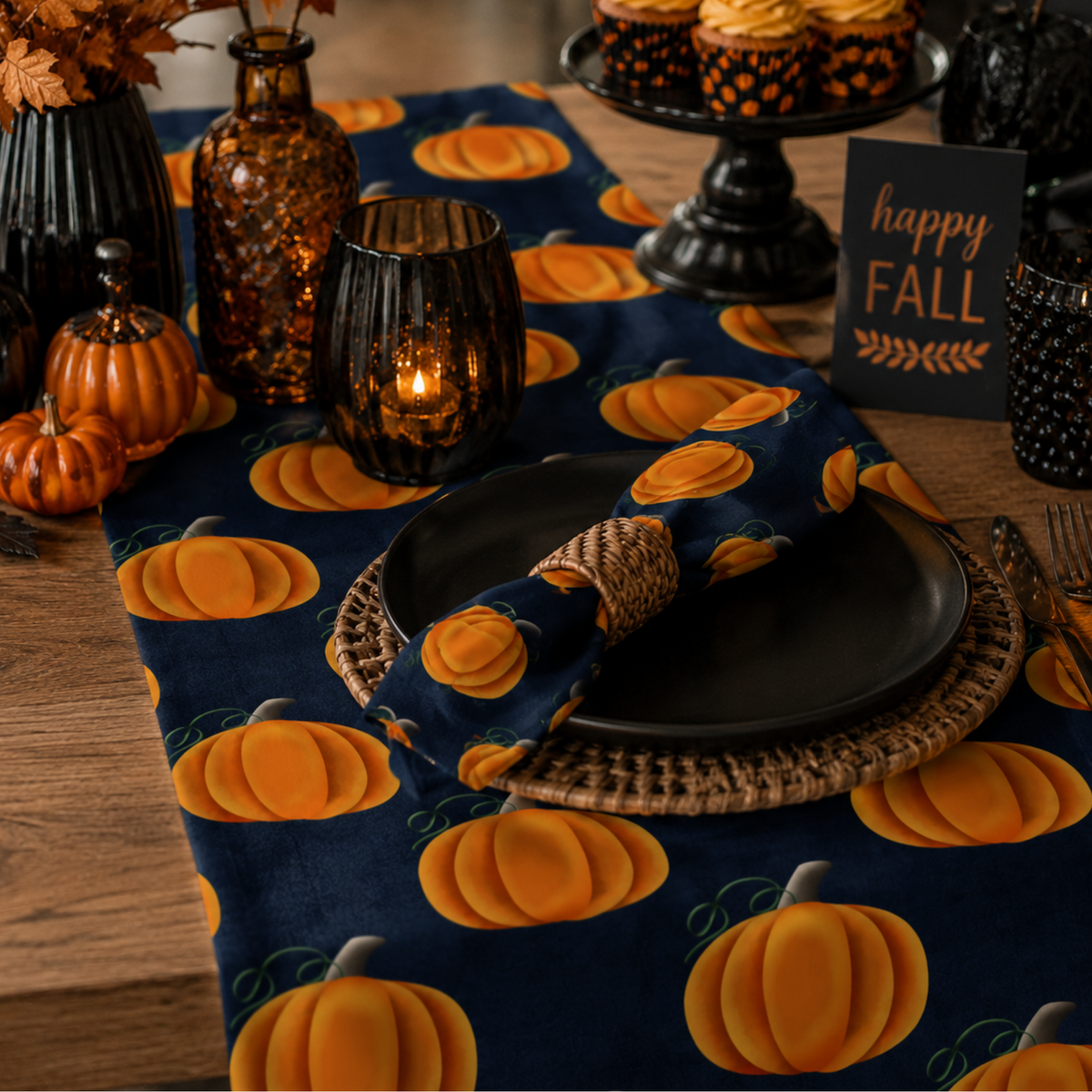 A styled autumn table setting featuring a navy blue table runner and matching napkin with a bright orange pumpkin print, set on a wooden table with black plates, amber glass candle holders, and a 'happy FALL' sign.